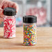 A hand holding a 3.5 oz. round plastic spice container full of red and white sprinkles.