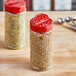 Two round plastic spice containers with red dual flapper lids with 7 holes on a table.