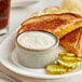 A grilled cheese sandwich with pickles and a cup of Ken's Foods Dill Pickle Aioli Sauce on a plate.