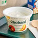 A white Chobani yogurt container on a table with a wooden spoon on top.