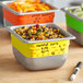 A group of Vigor 1/6 size food containers on a table with yellow write-on labels.