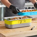 A man in black gloves writing on a yellow Vigor food pan on a counter.