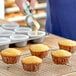 Corn muffins sit on a wire cooling rack. A person is pouring corn muffin batter into a muffin tin in the background.