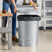 A man opening a Behrens galvanized steel trash can with a lid.