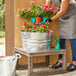 A person wearing gloves waters potted flowers in a Behrens galvanized steel tub on a patio table.