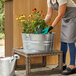 A person wearing gloves and holding a potted plant over a Behrens galvanized steel tub on a patio bench.