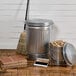 A Behrens galvanized steel trash can with an infinity lid and broom on a wood floor.