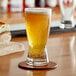 A glass of beer on a coaster on a wooden table next to a sandwich.