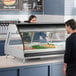 A woman putting food in a stainless steel Alto-Shaam countertop heated display case.