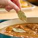 A hand holding a Hometown Provisions dried bay leaf over a bowl of soup.