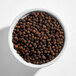 A white bowl filled with whole black peppercorns.