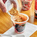 A hand pouring granola into a Del Monte Fruit Crunch Strawberry Non-Dairy Parfait.