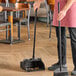 A person using an Advantage Supplies black open-lid dustpan to sweep a wood floor.