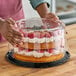 A person holding a D&W Fine Pack cake with a clear plastic scalloped lid over strawberries and whipped cream.