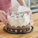 A person holding a cake in a D&W Fine Pack plastic container with a clear scalloped dome lid.