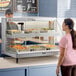 A woman in a pink shirt standing in front of a Hatco heated display case full of food.