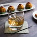 A Reserve by Libbey Symmetry rocks glass of liquid with ice on a table next to appetizers.