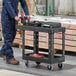 A man in a blue jumpsuit pushing a black Rubbermaid utility cart with tools on it.