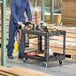 A man in blue overalls pushing a Rubbermaid utility cart with tools.