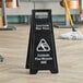 A close-up of a black Lavex "CAUTION Wet Floor" sign on a wood floor.