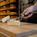 A person using a Boska stainless steel cheese knife to cut a piece of cheese on a wooden cutting board.