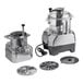 A commercial food processor with a clear plastic bowl, continuous feed attachment, and multiple metal discs.