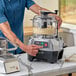 A commercial food processor with a clear plastic bowl and digital controls being operated on a kitchen counter.