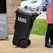 A black Lavex Pro 35-gallon wheeled rectangular trash can with a lid being moved by a person.