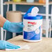 A gloved hand holding a white and blue container of WypAll CriticalClean disinfecting wipes with a blue lid.