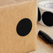 A brown box with a black circle on it next to a roll of tape with a Lavex black semi-gloss round inventory label on it.