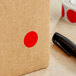 A brown box with a red circle on it next to a roll of Lavex Standard Red Semi-Gloss Paper Inventory Labels.