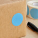 A brown box with a blue circle sticker next to a roll of tape.