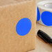 A brown box with a blue circle sticker next to a roll of Lavex Light Blue Semi-Gloss Paper Inventory Labels.