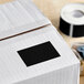 A white box with a black rectangular label on it. A roll of black rectangular labels.