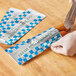 A person wearing gloves wrapping a hot dog in a Bagcraft foil wrapper with the words "Fresh and Delicious" and a honeycomb pattern.