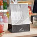 A white Bagcraft paper meal bag with a printed design and tin tie closure on a counter.