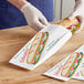 A person wearing white gloves cutting a sandwich into a Bagcraft Submarine sandwich bag.