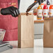 A person in black gloves using a metal scoop to pour coffee beans into a natural Kraft paper-lined bag with a tin tie closure.