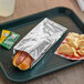 A hot dog in a Bagcraft silver foil wrapper on a tray.