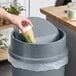 A hand putting a coffee cup into a Lavex swing-top wastebasket.