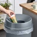 A hand pouring coffee from a paper cup into a Lavex round funnel wastebasket with a plant in the background.