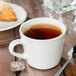 A Libbey Farmhouse porcelain cup with cream tea on a wooden table with a spoon and pastry.