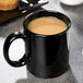 A Libbey black porcelain mug filled with brown liquid sits on a table next to a muffin.