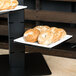 A white wide rim square porcelain plate displaying several bagels on a tiered stand.