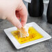 A person holding a piece of bread and dipping it into oil on a Libbey white square porcelain plate.