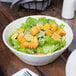 A Libbey round white porcelain serving bowl filled with salad, croutons, and cheese on a table.
