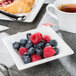 A Libbey ultra bright white square porcelain fruit bowl filled with blueberries and raspberries on a table with a spoon and a glass.