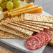 A plate with Clown Everything-Seasoned Flatbread Crackers, cheese, grapes, and sausage on a table in a deli.