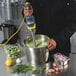 A person using a Hamilton Beach Big Rig immersion blender to mix green soup in a metal pot.