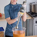 A woman using a Hamilton Beach BigRig immersion blender with orange liquid in a kitchen.